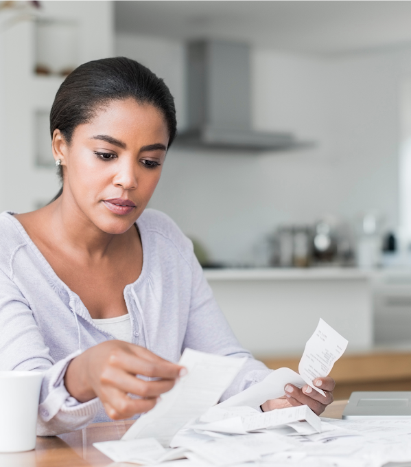 Une femme assise à une table fait du tri dans une pile de reçus. Un ordinateur portable apparaît à gauche de la pile et un mug à droite.