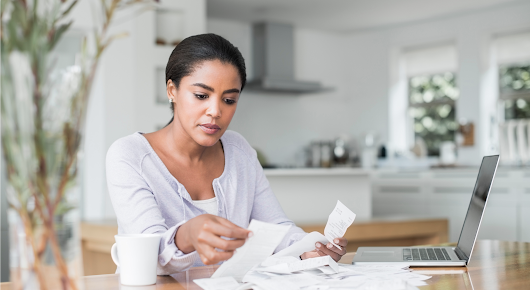 Une femme assise à une table fait du tri dans une pile de reçus. Un ordinateur portable apparaît à gauche de la pile et un mug à droite.