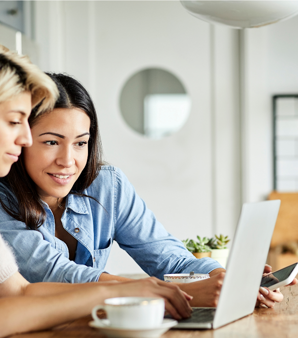 Deux femmes utilisent un ordinateur portable. L’une d’elles, portant un haut blanc, tape sur les touches du clavier, tandis que l’autre, portant un haut bleu, regarde l’écran de l’ordinateur un smartphone à la main.
