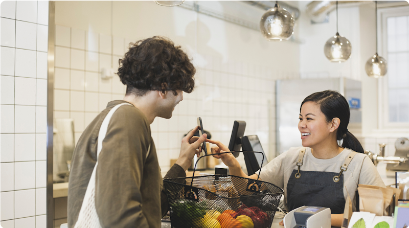 Un jeune homme utilise le paiement mobile pour acheter les produits qu’il a mis dans son panier. La caissière touche son téléphone en souriant.